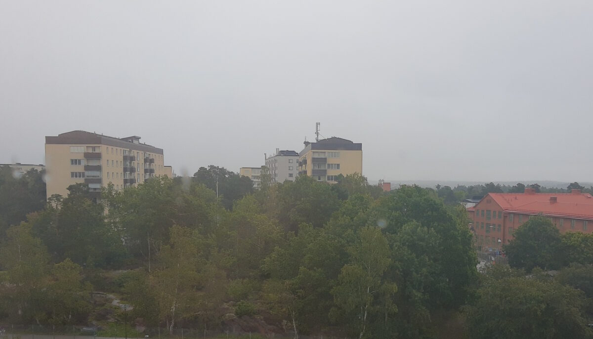 Ganska ljust grå himmel över gröna lövträd och höghus.